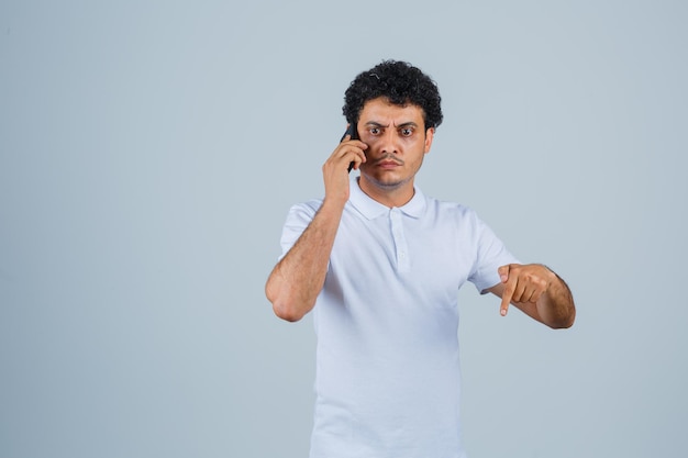 Junger Mann im weißen T-Shirt, der am Handy spricht, nach unten zeigt und nervös aussieht, Vorderansicht.