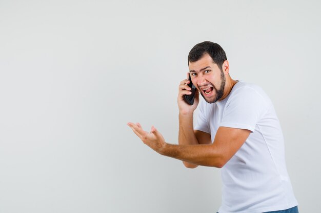 Kostenloses Foto junger mann im weißen t-shirt, der schreit, während er am telefon spricht und wütend, vorderansicht schaut. platz für text