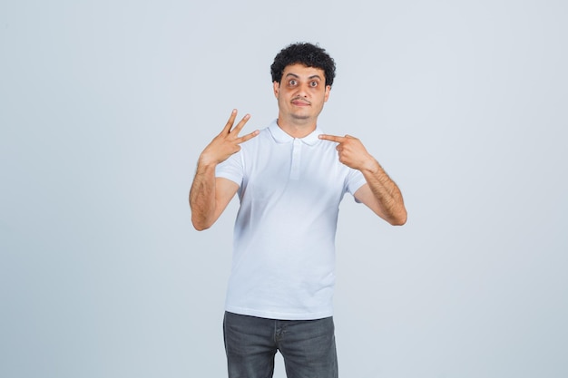 Junger Mann im weißen T-Shirt, Hose zeigt auf Nummer drei und sieht selbstbewusst aus, Vorderansicht.