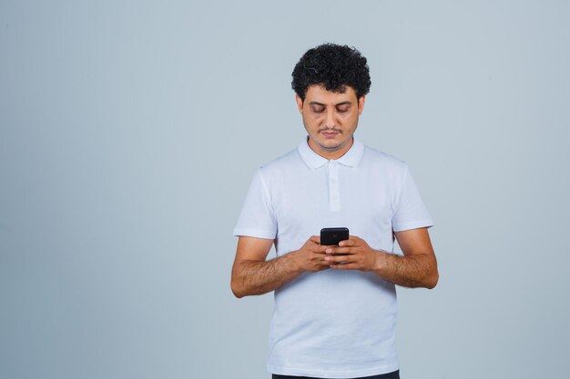 Junger Mann im weißen T-Shirt mit Handy und beschäftigt, Vorderansicht.