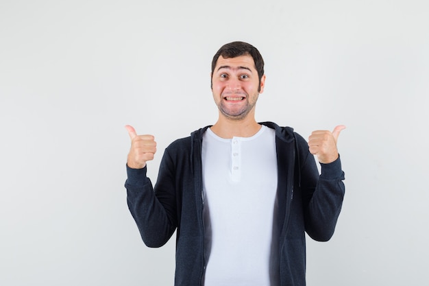 Junger Mann im weißen T-Shirt und im schwarzen Kapuzenpullover mit Reißverschluss vorne, der doppelte Daumen hoch zeigt und optimistisch, Vorderansicht schaut.