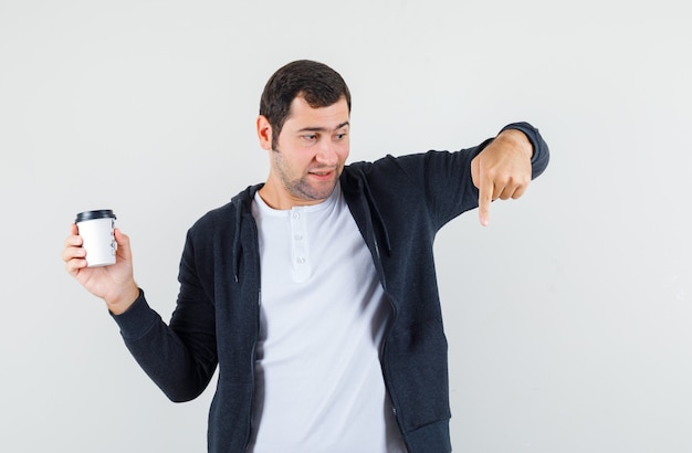 Junger Mann im weißen T-Shirt und im schwarzen Kapuzenpullover mit Reißverschluss vorne, der Kaffeetasse zum Mitnehmen hält und mit dem Zeigefinger nach unten zeigt und überrascht sieht, Vorderansicht.