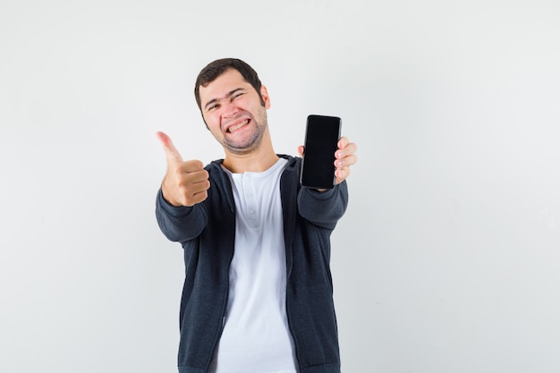Junger Mann im weißen T-Shirt und im schwarzen Kapuzenpullover mit Reißverschluss vorne, der Smartphone hält, zeigt Daumen hoch und schaut glücklich, Vorderansicht.