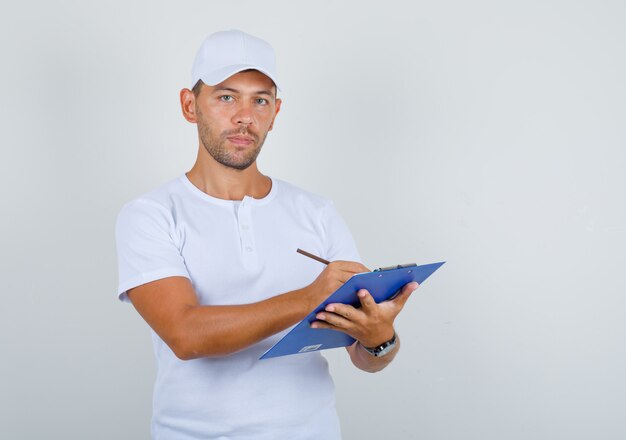 Junger Mann im weißen T-Shirt und in der Kappe, die Notizen auf Zwischenablage, Vorderansicht machen.