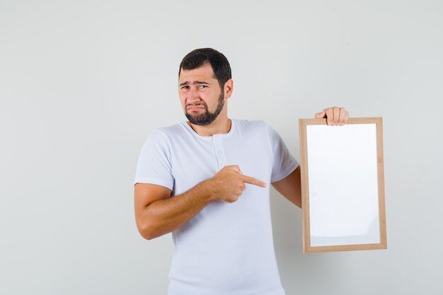 Junger Mann im weißen T-Shirt zeigt auf weiße Tafel und schaut unzufrieden, Vorderansicht.