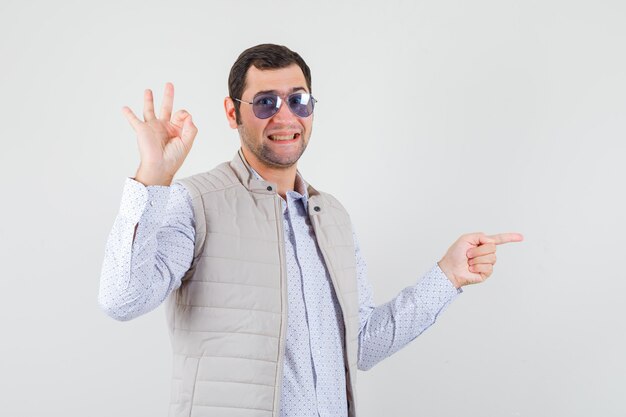 Junger Mann in beige Jacke und Kappe setzt Brille auf, während er mit Zeigefinger nach rechts zeigt und ok Zeichen zeigt und optimistisch, Vorderansicht schaut.