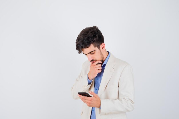 Junger Mann in blauem T-Shirt und weißer Anzugjacke, der Smartphone hält und in Denkpose steht und nachdenklich aussieht
