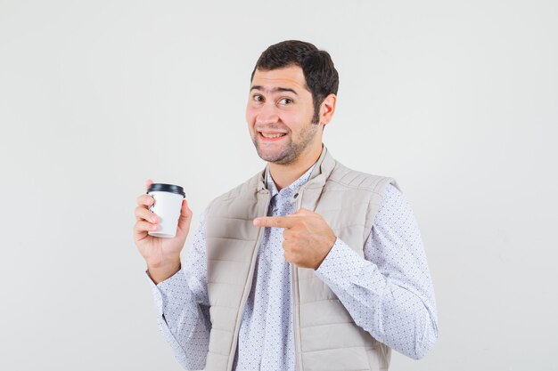 Junger Mann in der beigen Jacke, die auf Tasse Kaffee zum Mitnehmen mit Zeigefinger zeigt und optimistisch, Vorderansicht schaut.