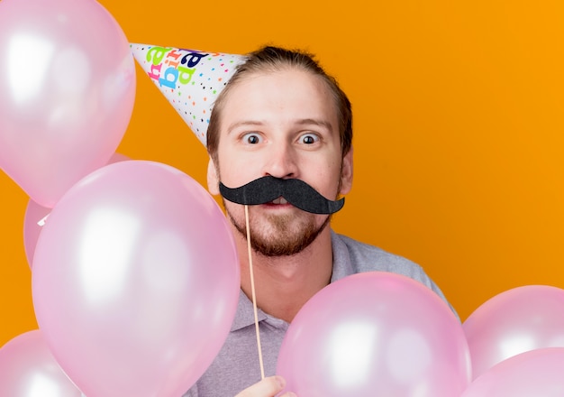 Kostenloses Foto junger mann in der feiertagskappe, die schnurrbart-party-stick hält, überraschte geburtstagsfeierkonzept, das mit luftballons über orange wand steht