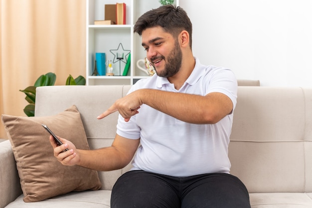 Junger mann in der freizeitkleidung, die smartphone hält, das mit zeigefinger glücklich und positiv auf einer couch im hellen wohnzimmer sitzend zeigt