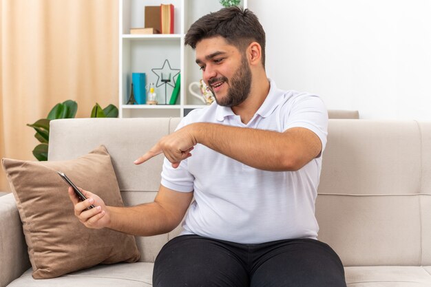 Junger Mann in der Freizeitkleidung, die Smartphone hält, das mit Zeigefinger glücklich und positiv auf einer Couch im hellen Wohnzimmer sitzend zeigt