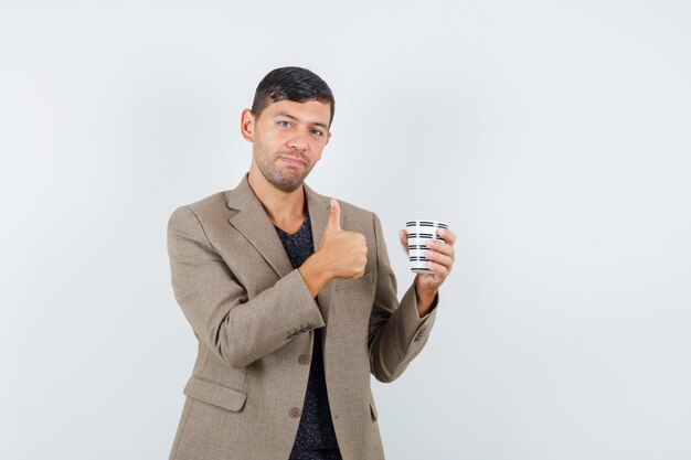 Junger Mann in der graubraunen Jacke, die Tasse hält, während Daumen oben zeigt und erfreut, Vorderansicht schaut.