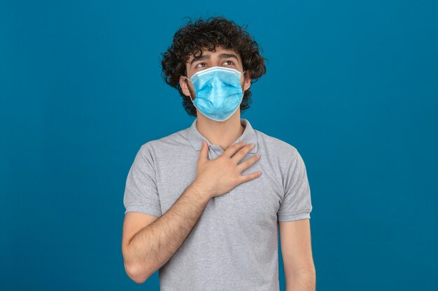 Junger Mann in der medizinischen Schutzmaske, die auf Brust berührt, um Lunge beim Atmen über isoliertem blauem Hintergrund zu überprüfen
