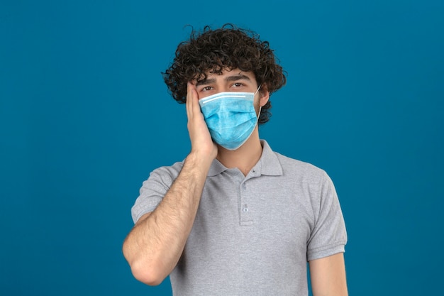 Junger Mann in der medizinischen Schutzmaske, die den Kopf berührt, der krank schaut und Kopfschmerzen hat, die mit unglücklichem Gesicht über lokalisiertem blauem Hintergrund stehen