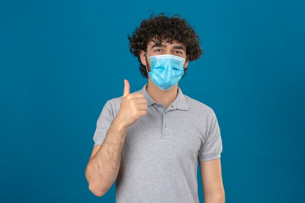 Junger Mann in der medizinischen Schutzmaske, die Kamera mit glücklichem Gesicht betrachtet, das Daumen oben über lokalisiertem blauem Hintergrund zeigt