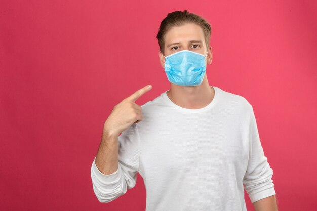 Junger Mann in der medizinischen Schutzmaske, die mit Finger auf sein Gesicht zeigt und Kamera über lokalisiertem rosa Hintergrund betrachtet