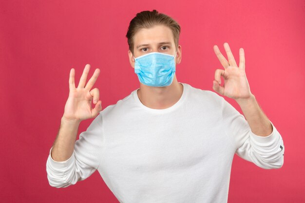 Junger Mann in der medizinischen Schutzmaske, die ok Zeichen mit den Fingern und den Händen betrachtet Kamera über lokalisiertem rosa Hintergrund zeigt