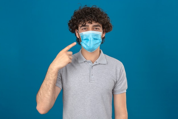 Junger Mann in der medizinischen Schutzmaske zeigt auf seine Maske, die Kamera mit Lächeln über lokalisiertem blauem Hintergrund betrachtet