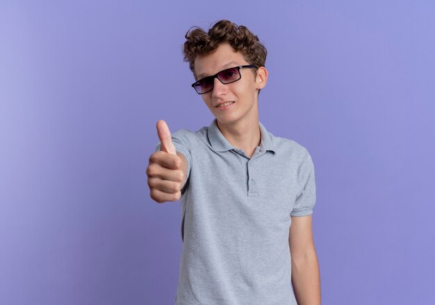Junger Mann in der schwarzen Brille, die das graue Poloshirt lächelnd zeigt, zeigt Daumen hoch stehend über blauer Wand