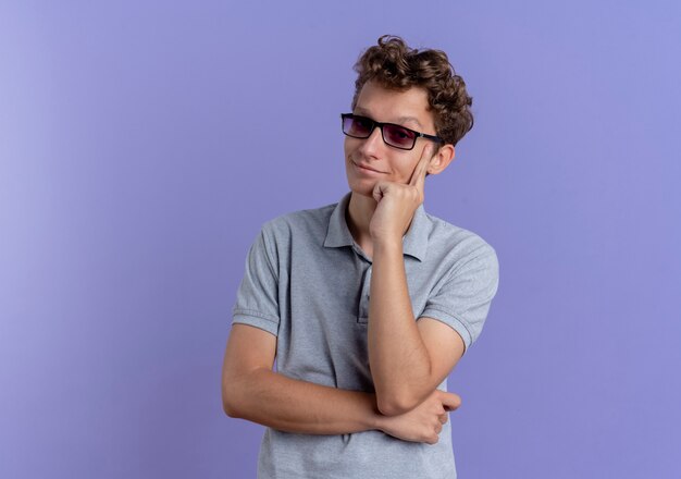 Junger Mann in der schwarzen Brille, die das graue Poloshirt trägt, das beiseite lächelnd zuversichtlich über der blauen Wand steht