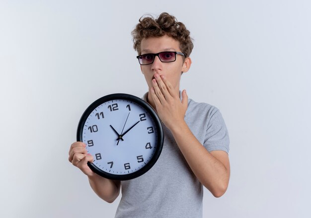 Junger Mann in der schwarzen Brille, die das graue Poloshirt trägt, das Wanduhr hält, die Mund mit Hand bedeckt, die über weißer Wand schockiert steht