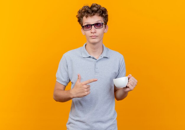 Junger Mann in der schwarzen Brille, die graues Poloshirt hält, das Kaffeetasse hält, die mit dem Finger darauf zeigt und mit ernstem Gesicht über Grün beiseite schaut