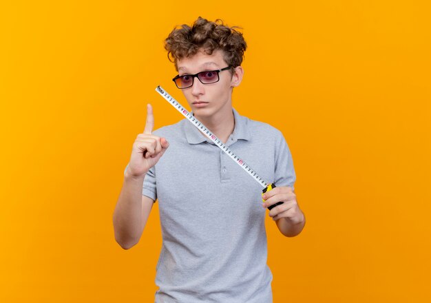 Junger Mann in der schwarzen Brille, die graues Poloshirt hält, das Maßband lächelnd glücklich und positiv zeigt Zeigefinger mit neuer Idee über Orange