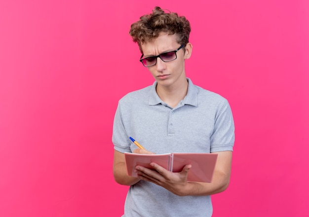 Junger Mann in der schwarzen Brille, die graues Poloshirt hält, das Notizbuch mit Stift hält, der es mit ernstem Stirnrunzeln Gesicht über Rosa betrachtet