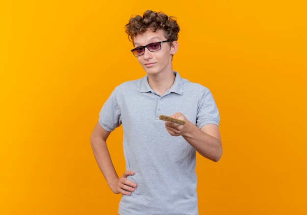 Junger Mann in der schwarzen Brille, die graues Poloshirt hält, das Pinselangebot an der Kamera über Orange hält