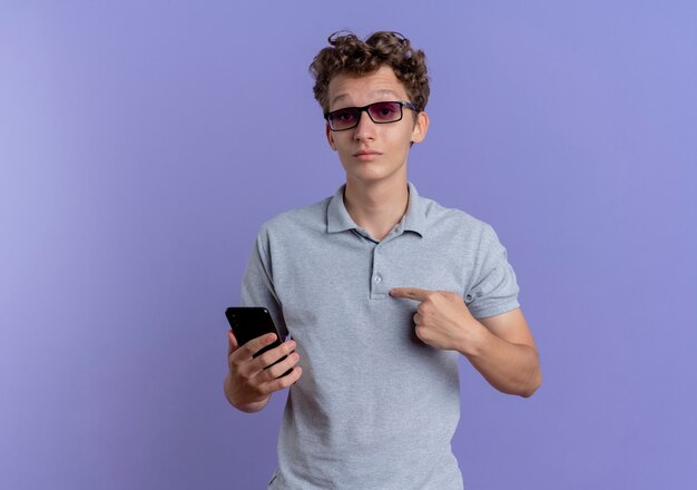 Junger Mann in der schwarzen Brille, die graues Poloshirt hält, das Smartphone zeigt, das auf sich zeigt, verwirrt über der blauen Wand stehend