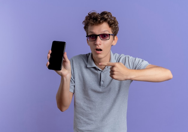 Junger Mann in der schwarzen Brille, die graues Poloshirt hält, das Smartphone zeigt, das mit Zeigefinger darauf zeigt, dass es überrascht ist, über blauer Wand zu stehen