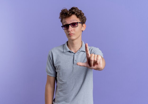 Junger Mann in der schwarzen Brille, die graues Poloshirt mit ernstem Gesicht zeigt Zeigefingerwarnung steht über blauer Wand