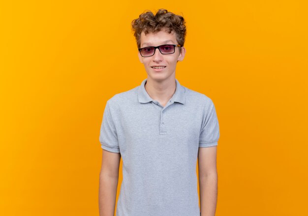 Junger Mann in der schwarzen Brille, die graues Poloshirt mit Lächeln auf Gesicht über Orange trägt