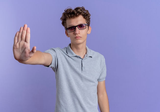 Junger mann in der schwarzen brille, die graues poloshirt mit stirnrunzelndem gesicht trägt, das stoppgeste mit hand steht, die über blauer wand steht