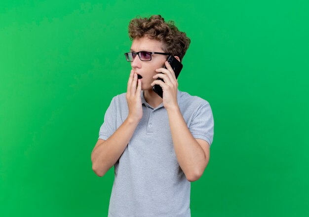 Junger Mann in der schwarzen Brille, die graues Poloshirt trägt, das auf Handy spricht, das über Grün erstaunt und überrascht wird