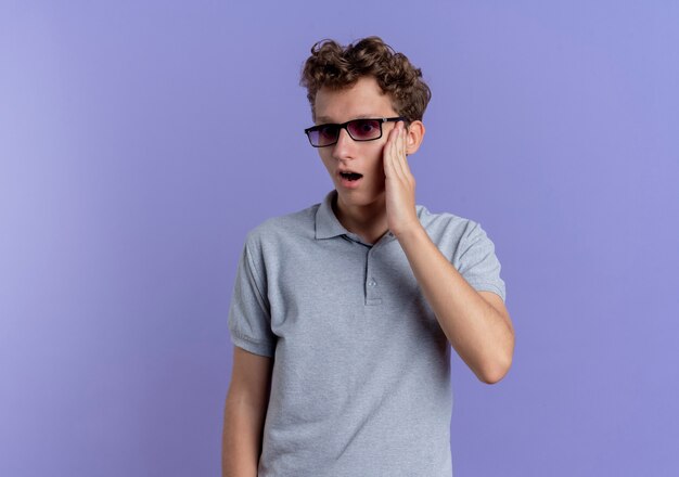 Junger Mann in der schwarzen Brille, die graues Poloshirt trägt, das beiseite schaut, verwirrt und besorgt steht über blauer Wand