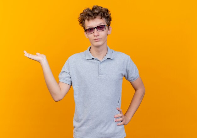 Junger Mann in der schwarzen Brille, die graues Poloshirt trägt, das etwas mit Arm der Hand darstellt, die über Orange zuversichtlich schaut