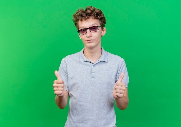 Junger Mann in der schwarzen Brille, die graues Poloshirt trägt, das hapy und positiv lächelnd zeigt Daumen hoch, die über grüner Wand stehen