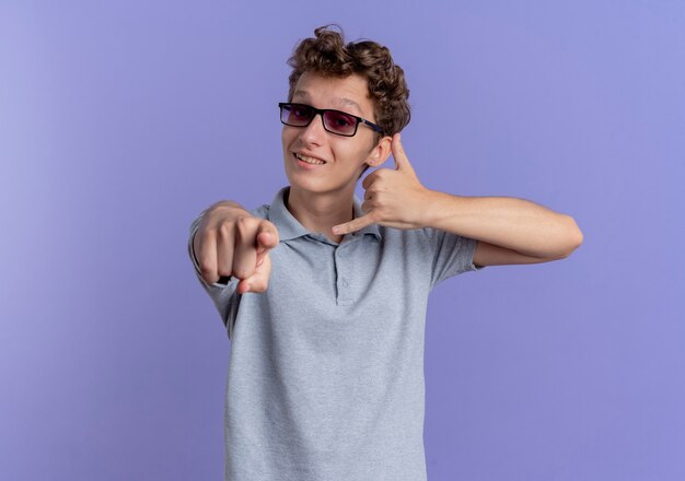 Junger Mann in der schwarzen Brille, die graues Poloshirt trägt, das mit dem lächelnden Finger zeigt, der mich Geste nennt, die über blaue Wand steht