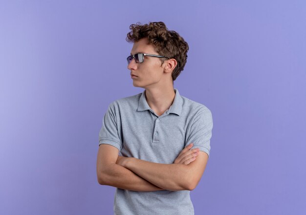 Junger Mann in der schwarzen Brille, die graues Poloshirt trägt, das mit ernstem Gesicht mit gekreuzten Händen auf Brust über Blau beiseite schaut