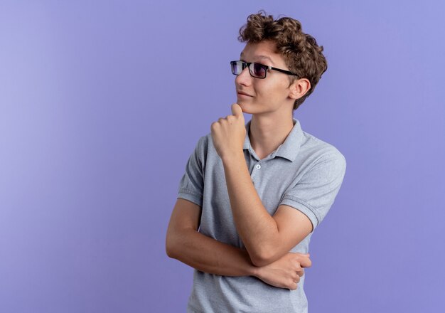 Junger Mann in der schwarzen Brille, die graues Poloshirt trägt, das mit Hand auf Kinn betrachtet, das über Blau denkt