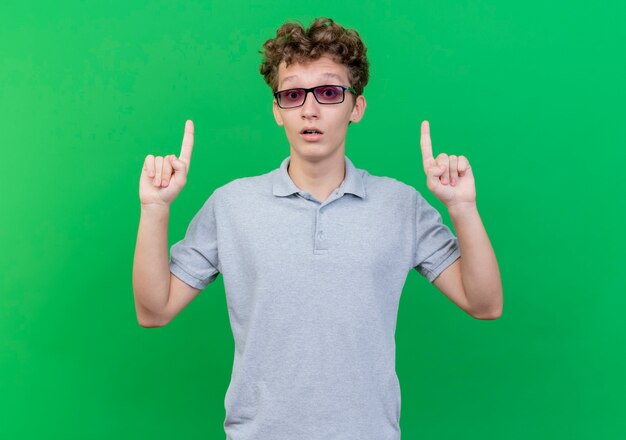 Junger Mann in der schwarzen Brille, die graues Poloshirt trägt, das mit idex Fingern oben zeigt, überrascht über grüner Wand stehend