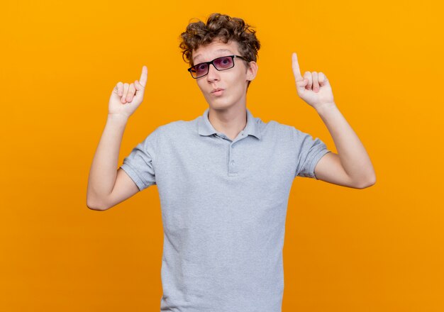 Junger Mann in der schwarzen Brille, die graues Poloshirt trägt, das mit idex Fingern oben zeigt, überrascht, über orange Wand stehend