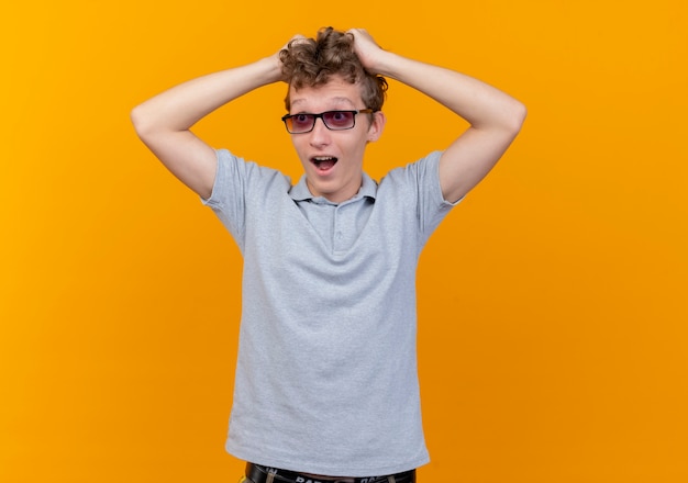 Junger Mann in der schwarzen Brille, die graues Poloshirt trägt, das seinen Kopf berührt, der über Orange überrascht und aufgeregt ist