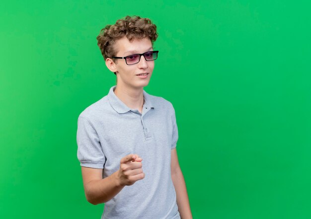 Junger Mann in der schwarzen Brille, die graues Poloshirt trägt, das sicher zeigt, mit Zeigefinger auf Kamera über Grün zu zeigen