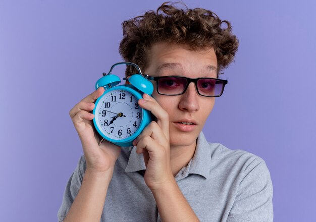 Junger Mann in der schwarzen Brille, die graues Poloshirt trägt, das Wecker zeigt, der verwirrt über blauer Wand steht