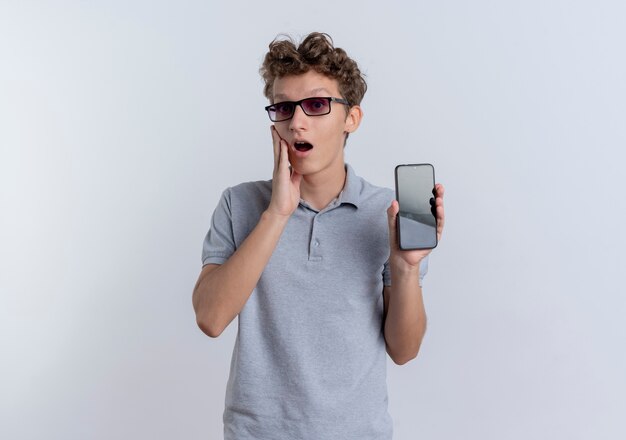 Junger Mann in der schwarzen Brille, die graues Poloshirt trägt Smartphone zeigt, das überrascht und erstaunt steht, das über weißer Wand steht