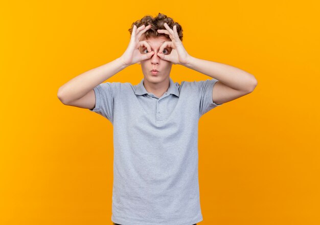 Junger Mann in der schwarzen Brille, die graues Poloshirt trägt und binokulare Geste mit Fingern macht, die durch Finger über Orange schauen