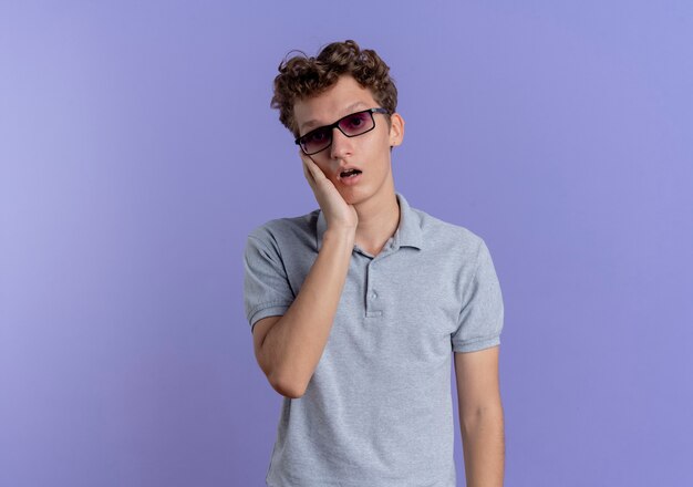 Junger Mann in der schwarzen Brille, die verwirrtes und sehr besorgtes graues Poloshirt trägt, das über blaue Wand steht