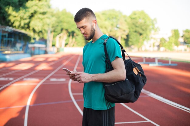 Junger Mann in drahtlosen Kopfhörern benutzt träumerisch sein Handy, während er Zeit auf der Laufstrecke des Stadions verbringt